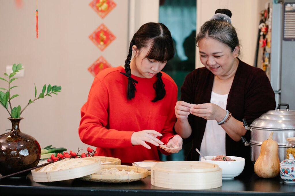 Cheerful Asian grandma with granddaughter filling dough while cooking dim sum at table with steamer and fresh squash